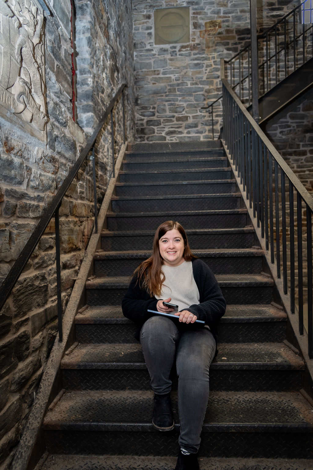 Erika, standing in a green business casual jumpsuit with a black cardigan, crossing her arms while smiling and looking at the camera. Behind her, various green bushes and a bed of grass with street lamps in the park during an overcast yet bright day.